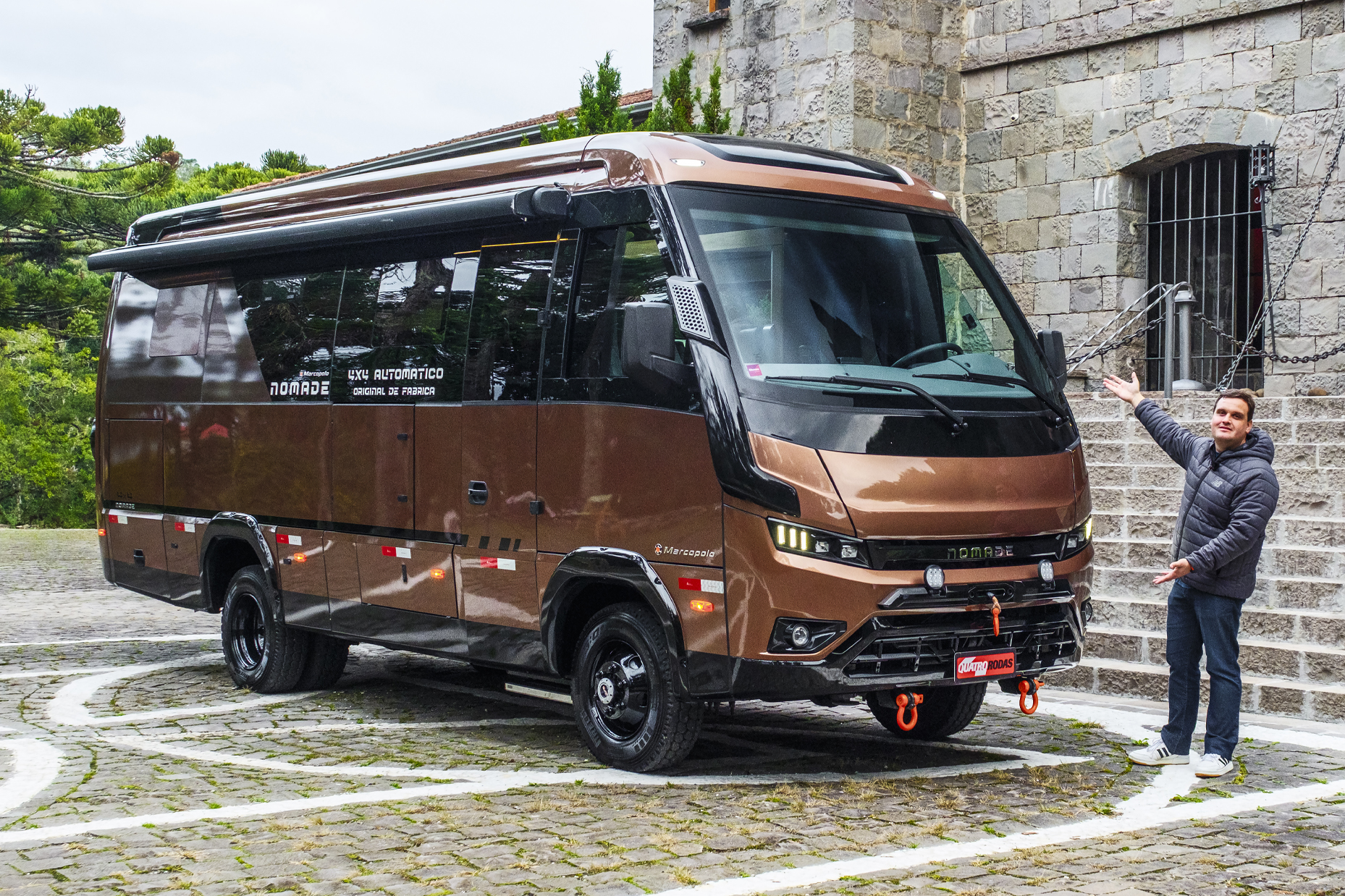 Homem sorridente de jaqueta cinza e calça jeans aponta para um motorhome marrom-bronze com toldo lateral estendido, estacionado em pátio de pedras com um prédio antigo ao fundo